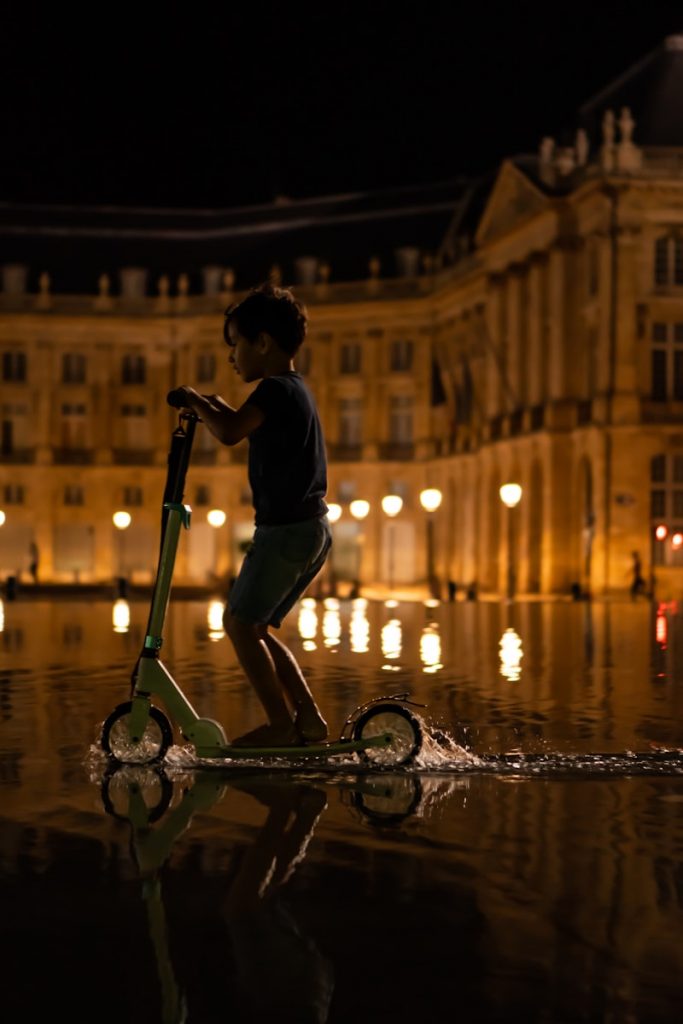 a boy on a scooter in the water with a building in the background